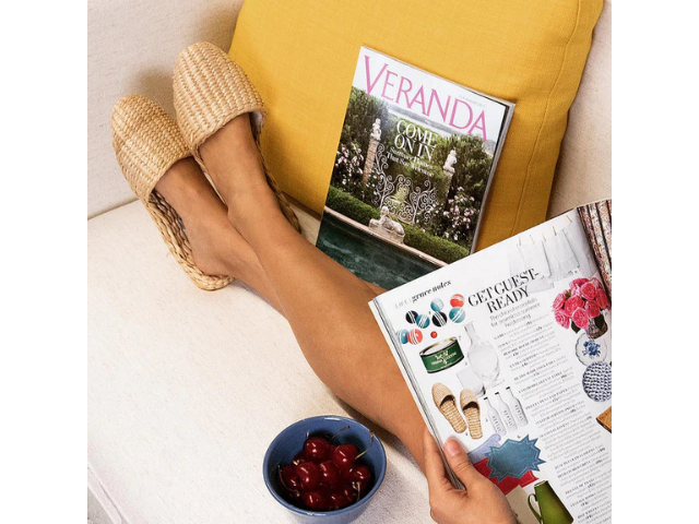 Woman's legs up on pillow while wearing straw slippers and looking at magazine