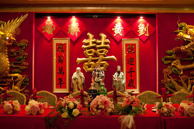 A large gold double happiness wall hanging with auspicious scrolls behind a table set with red tablecloth and flowers for bride, groom, and wedding party