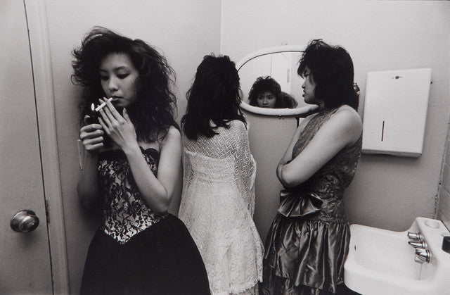 Three women in prom dresses in bathroom, one smoking