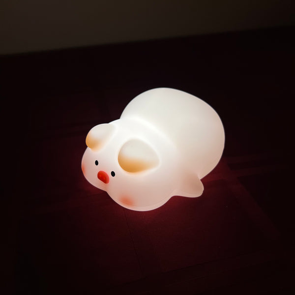 A small pig-shaped night light glowing with warm white light, featuring a cute face with black eyes, a red nose, and pink cheeks, resting on a dark surface.