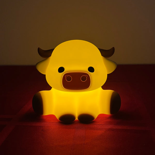 A glowing yellow cartoon cow night light with dark brown horns, snout, and feet, sitting on a red surface against a dark background.