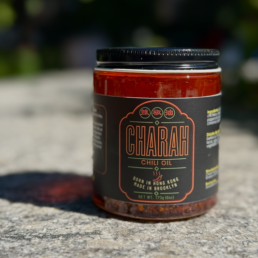 A jar of Charah Chili Oil sits on a textured stone surface outdoors, with a blurred natural background. The product features a black label with orange and white text, and a black metal lid.