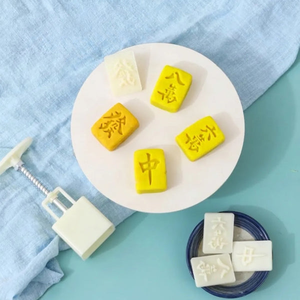 Five miniature yellow and white mahjong tile-shaped mooncakes arranged on a white plate, alongside a white tile-shaped mold and a blue-and-white lid, set against a light blue fabric and mint background.
