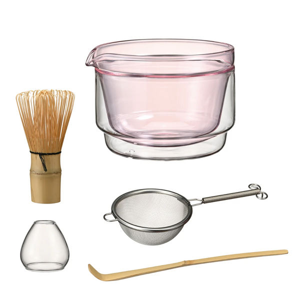 Set of tea-making tools including a pink glass container, whisk, small bowl, strainer, and wooden spoon on a white background.