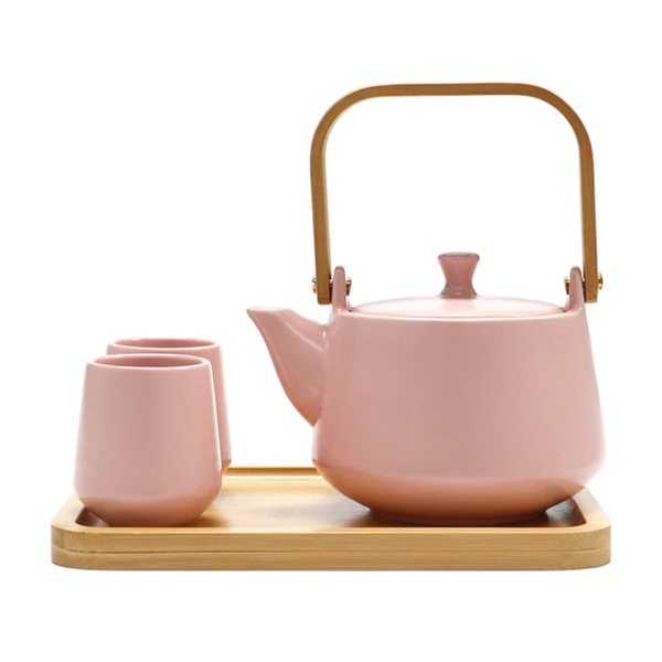 Pink ceramic teapot with wooden handle and two matching cups on a wooden tray against a white background