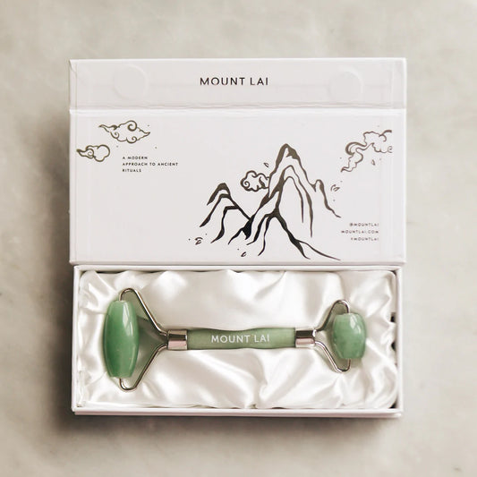 A Mount Lai jade facial roller presented in a white box with a drawing of mountains and the brand name. The roller has a green jade handle with a dual-sided design.