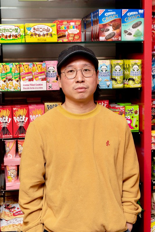 Andrew Yung wearing glasses and yellow sweatshirt in front of Asian snacks at Pearl River Mart