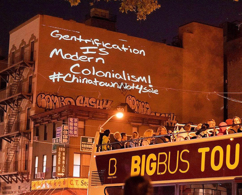 Light installation with text, "Gentrification is modern colonialism #chinatownartbrigade" on side of building with double decker tour bus in front