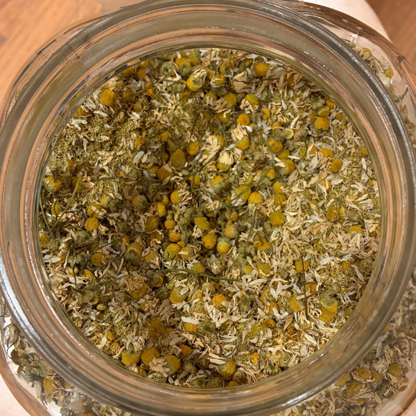 Top-down view of loose-leaf herbal tea or chamomile mixture in a clear glass jar, showing yellow-green flower heads mixed with dried green leaves and pale stems.