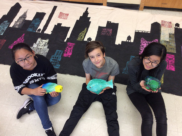 Students sitting on floor on top of city mural with papier mache fish 