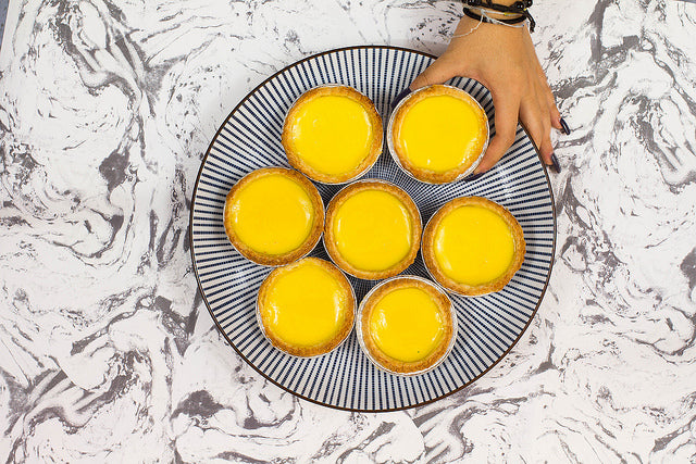 Blue and white striped dish with bright yellow egg tarts, and hand holding a tart 