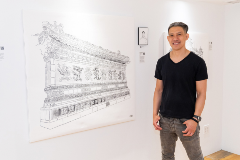 A man in a black v-neck t-shirt and gray jeans stands in a gallery next to a large black and white architectural sketch of the Terracotta Army on a white wall.