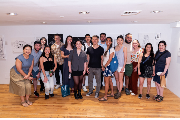 A group of twenty people posing together in a gallery or studio with white walls, wood floors, and framed artwork in the background.