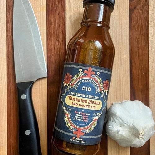 Bottle of Tamarind Heads BBQ sauce between a knife and garlic clove on a cutting board