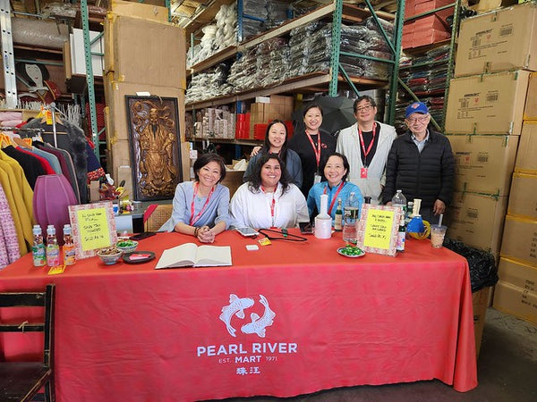 Seven Pearl River Mart employees behind table with red Pearl River Mart tablecloth in their Queens warehouse