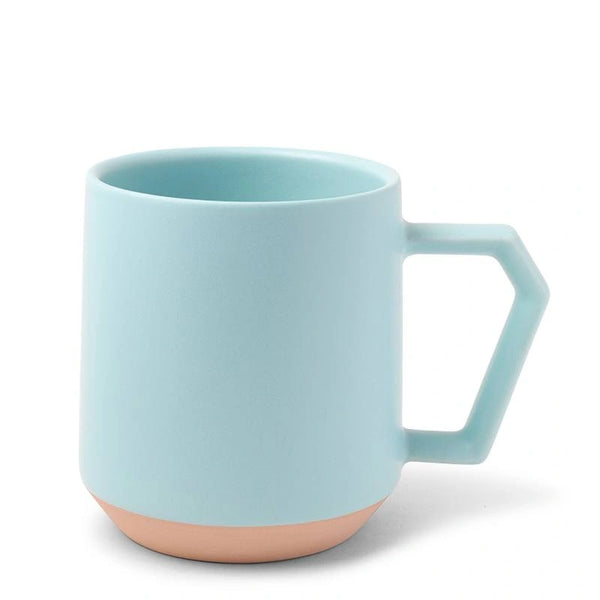 A light blue ceramic mug with an angular handle and a matte finish. The base features a contrasting unglazed terracotta brown. The tone is modern and minimalist.