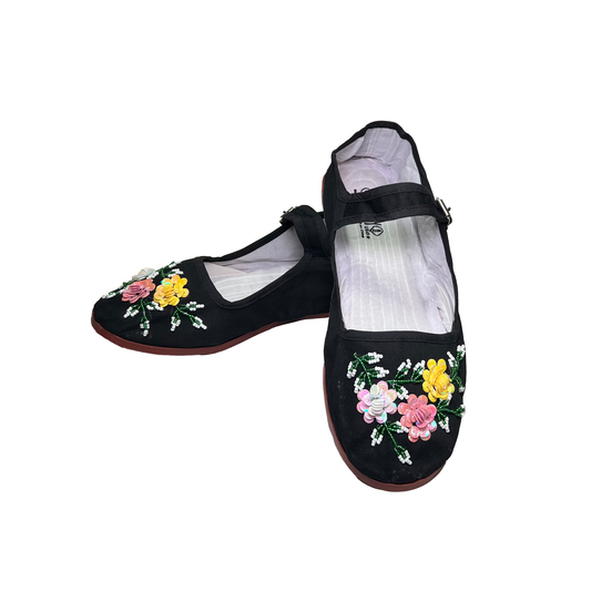 A pair of black flat shoes with traditional Chinese floral embroidery on the toes, featuring pink, yellow, and white flowers with green leaves, displayed on a solid white background.