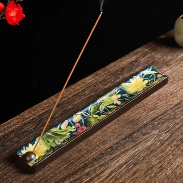 Enamel ceramic incense burner with lotus design on a blue-green background. A sample piece of incense is burning at an angle.