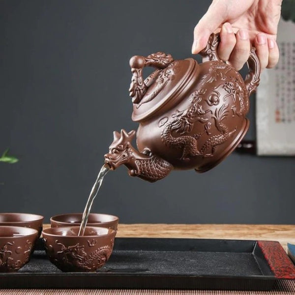 Natural Clay Dragon Tea Set with one teapot and four tea cups. A dragon design is carved on the the teapot spout, body, handle, lid, and tea cups. The teapot is being used to pour tea into a tea cup.