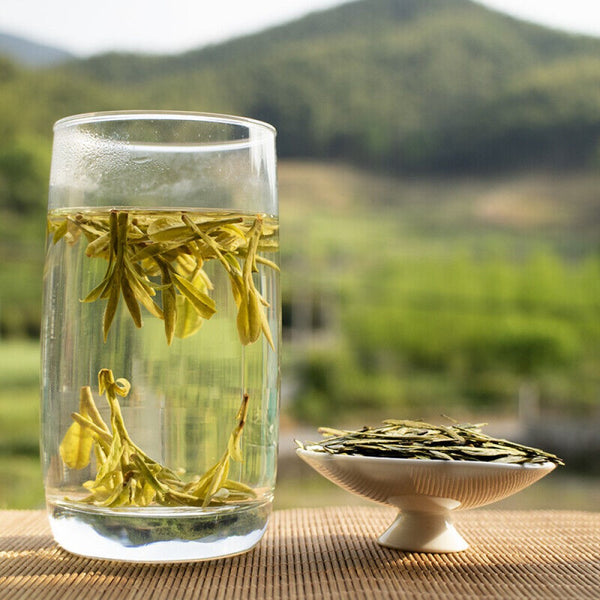 Ming Qing Longjing Green Tea brewed in a glass of water with a mini dish of loose tea leaves on the side.