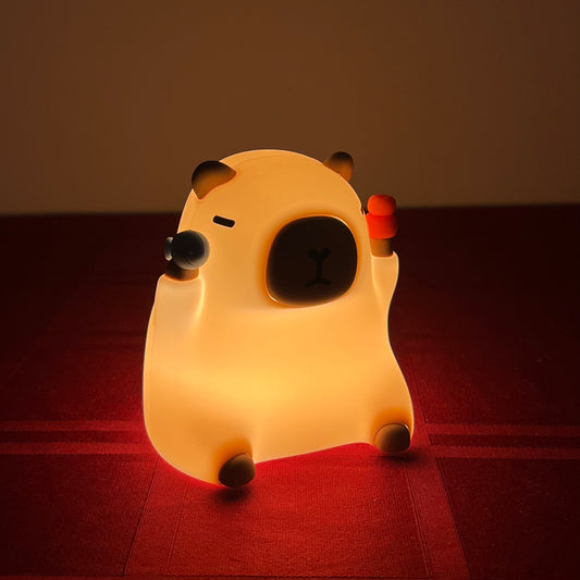 A cute, cartoonish capybara-shaped night light glowing with a warm yellow light, sitting on a dark red textured surface against a dark brown background.