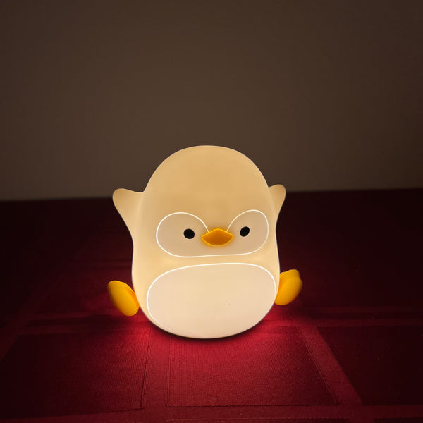 A cute penguin-shaped night light glowing with warm yellow and white light, resting on a dark red textured surface against a dark brown background.