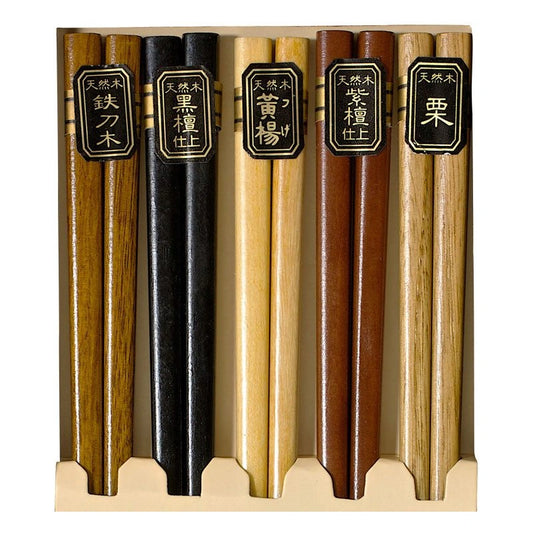 Five pairs of Japanese chopsticks in a wooden display box, each with a black label featuring gold Japanese characters indicating different wood types.