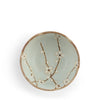 A top-down view of a ceramic bowl with a pale green glaze, decorated with hand-painted brown branches and white blossoms, on a white background.