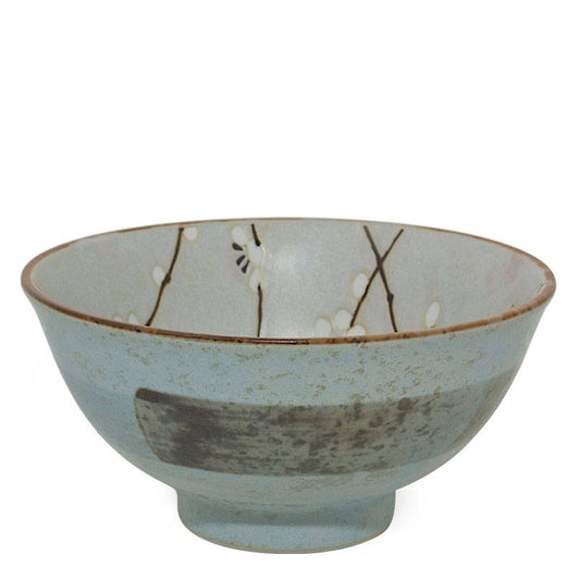 A handmade ceramic bowl with a light blue-gray glaze, featuring a rustic brown rim, a dark gray textured patch on the front, and delicate black branch and white blossom motifs painted inside.