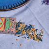 A colorful patterned tin of whole spices spilled onto a light blue fabric surface, with blue, yellow, and orange seeds scattered beside the open container.