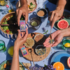 A top-down lifestyle shot of a table setting with multiple hands holding and smelling lavender sachets and loose-leaf lavender. The scene is vibrant and styled with fresh fruits, salads, and cups of tea on a blue tablecloth.
