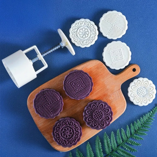 White plastic round mold set with four different embossed patterns, shown alongside four purple mooncakes on a wooden cutting board, on a blue background with a green fern leaf.