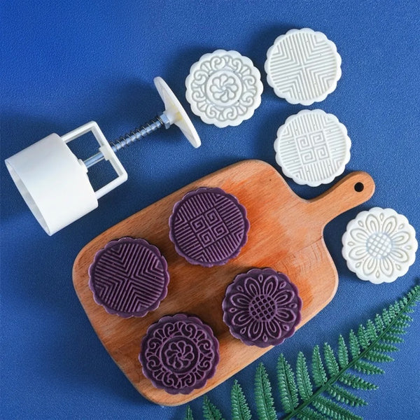 White plastic round mold set with four different embossed patterns, shown alongside four purple mooncakes on a wooden cutting board, on a blue background with a green fern leaf.