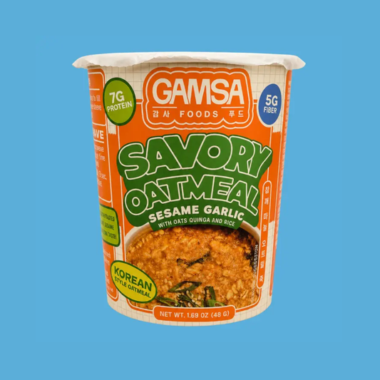 A cup of Gamsa Savory Oatmeal Sesame Garlic on a solid blue background. The packaging features orange and white colors with green text, and a photo of the prepared oatmeal with sesame garlic is shown on the front label.