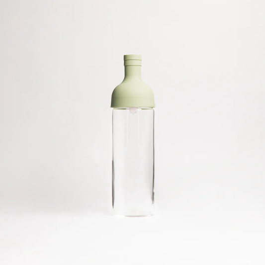 A clear glass bottle with a light sage green silicone cap, centered against a seamless white studio background.
