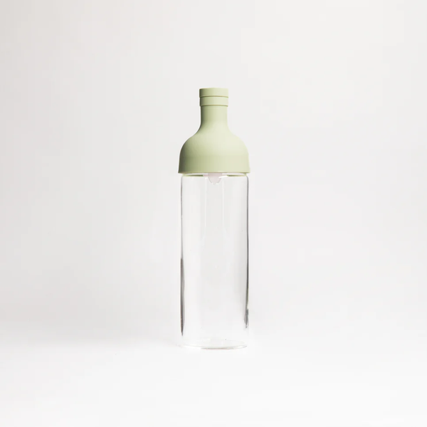 A clear glass bottle with a light sage green silicone cap, centered against a seamless white studio background.