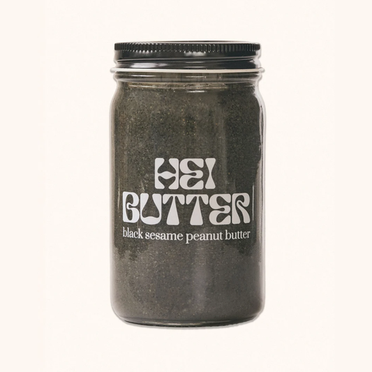A clear glass jar filled with dark black sesame peanut butter, featuring a black metal screw-on lid and white retro-style text that reads 'HEI BUTTER' and 'black sesame peanut butter' on the front, set against a plain off-white background.