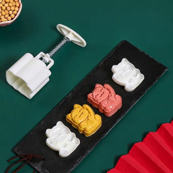 A white plastic cat-shaped stamp mold with a screw handle is shown beside a black serving tray holding four molded cat-shaped items in white, yellow, pink, and red, on a dark green background.