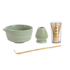 Tea-making set with green ceramic bowl, small container, wooden spoon, and transparent container with wooden sticks on a white background.