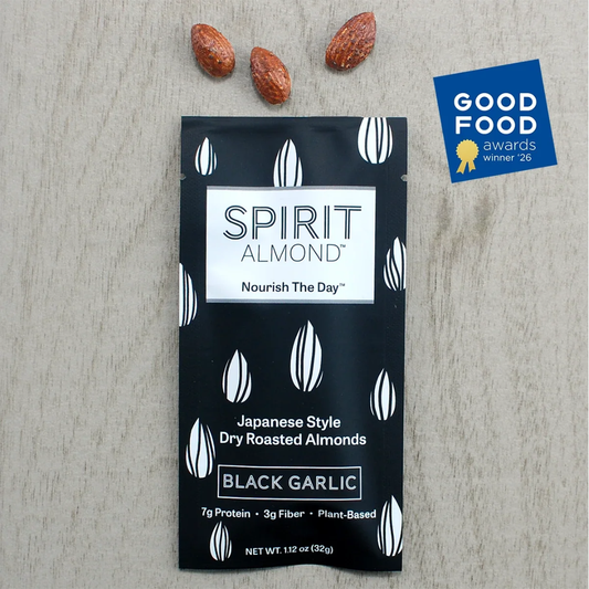 A single packet of Spirit Almond Black Garlic dry roasted almonds lies on a light wood surface, with three whole almonds placed above the package. A Good Food Awards Winner 26 badge appears in the upper right corner.