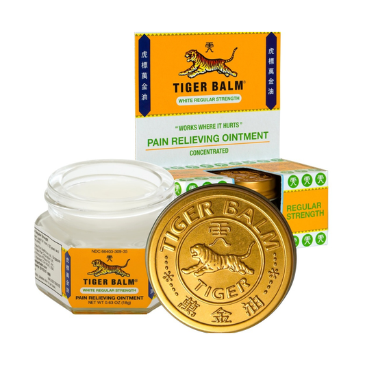 Tiger Balm white regular strength pain relieving ointment shown with its orange and white retail box, an open jar of white ointment, and a round gold balm disc in the foreground on a white background.