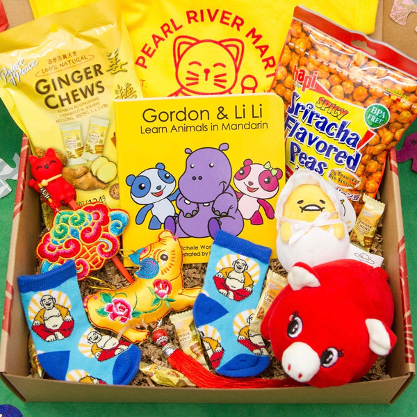 Gift box full of cute baby items, including yellow and red lucky cat onesie, a Gordon and Li Li Chinese book, a red plush pig, a Gudetama keychain, baby socks, a brocade pig ornament, and snacks for the parents. Please note substitutions might be made depending on availability.