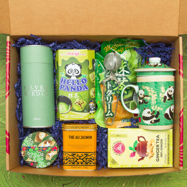 A box of tea lover items, including a canister of high quality green tea, matcha panda cookies, tin of jasmine tea, matcha candy, a mug, a handheld tea strainer, and ginger tea