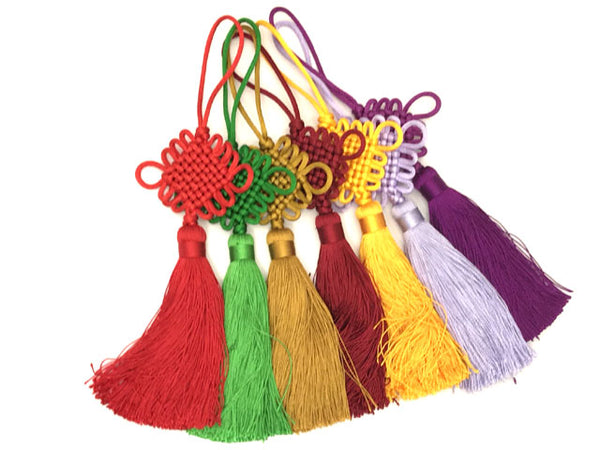 Six Chinese knot tassels next to each other. The colors from left to right are red, green, gold, burgundy, yellow, lavender and purple 