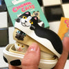 Chonk cats dolls book and box on a checkered surface in the background. While someone holds one cat doll nesting the other two