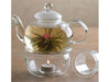 jasmine tea bud bloomed in tea pot glassware. Attached under is a glassware  holding a lit candle inside. All this resting on a table.